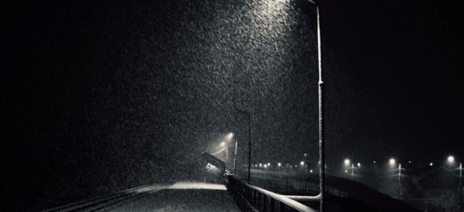lamp post at night in winter landscape with foosteps in the snow and train tracks on the left and distant lights on the right