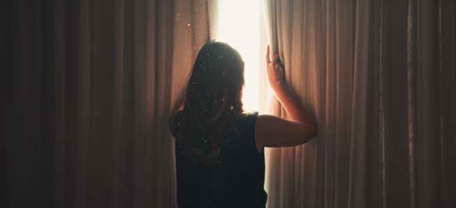 woman opening window curtain in shining daylight