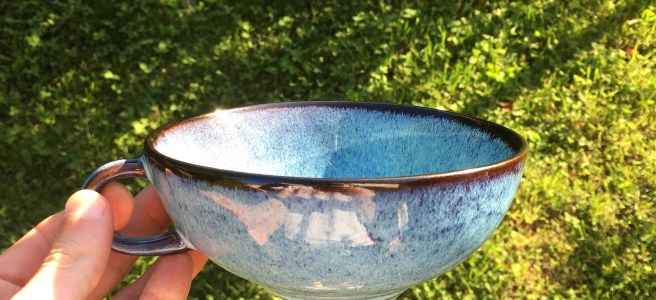 Chinese style cup of tea held above sunlit grass with left hand